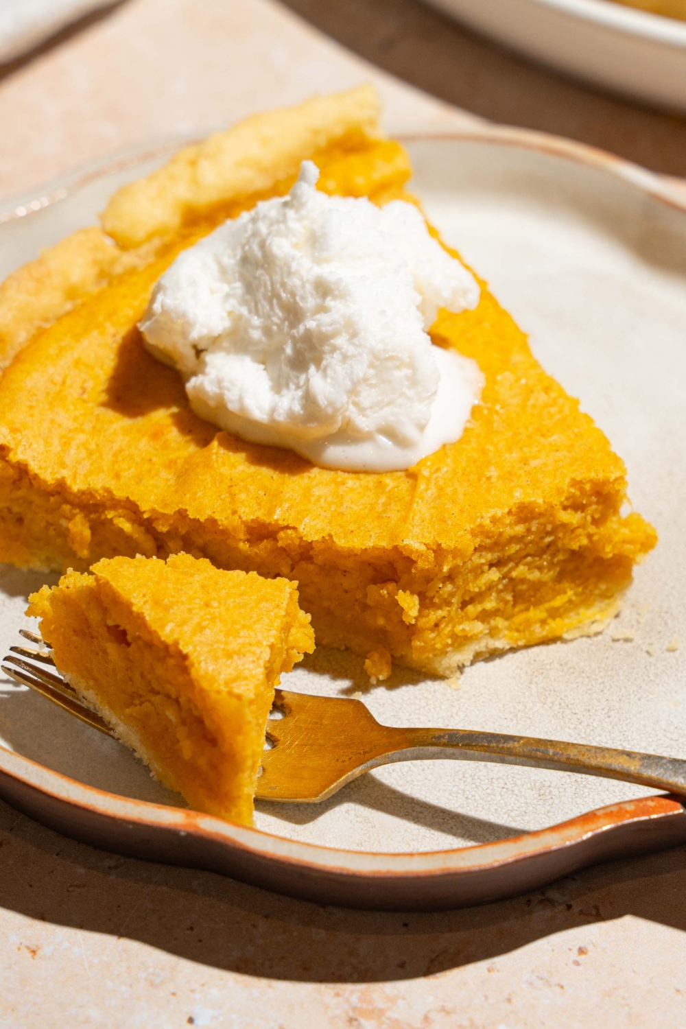 A plate with a slice of cream cheese pumpkin pie topped with a dollop of whipped cream. A fork is on the plate with a bite of pie. The plate is on a tan counter.