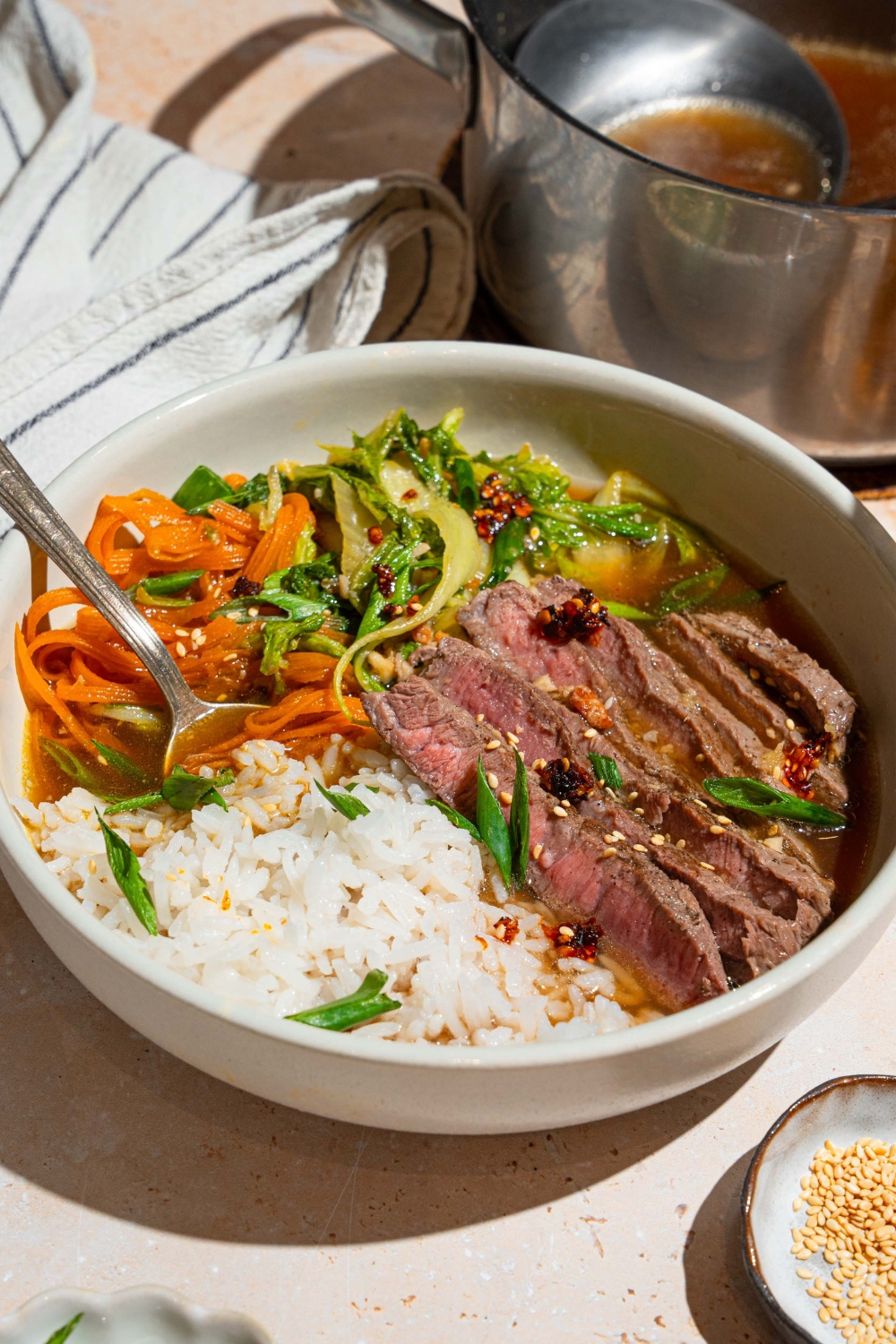 A steak and rice broth bowl with jasmine rice, sliced sirloin, shredded carrots and bok choy in a white bowl filled with broth.