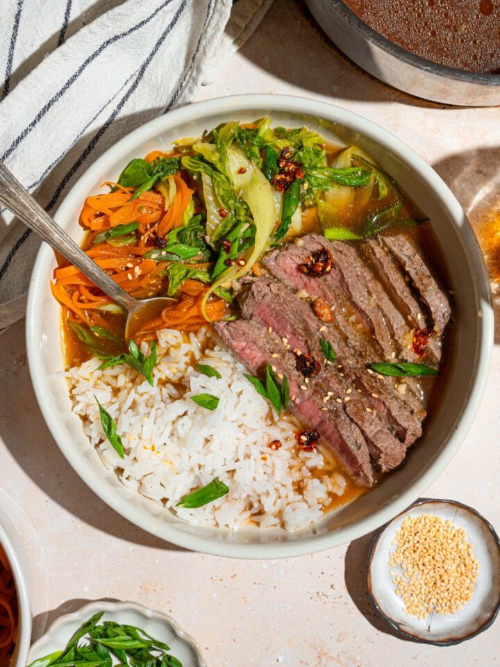 A brothy steak and rice bowl with sliced steak, shredded carrots and bok choy, and jasmine rice in broth in a white bowl.