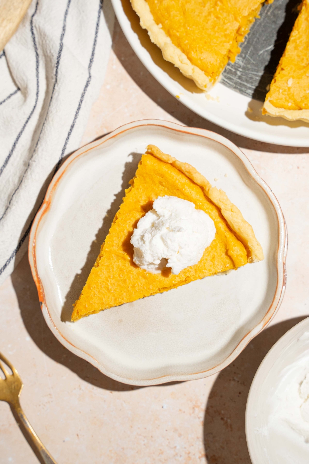 A plate with a slice of cream cheese pumpkin pie topped with a dollop of whipped cream. The plate is on a tan counter with a whole pie and white striped napkin.