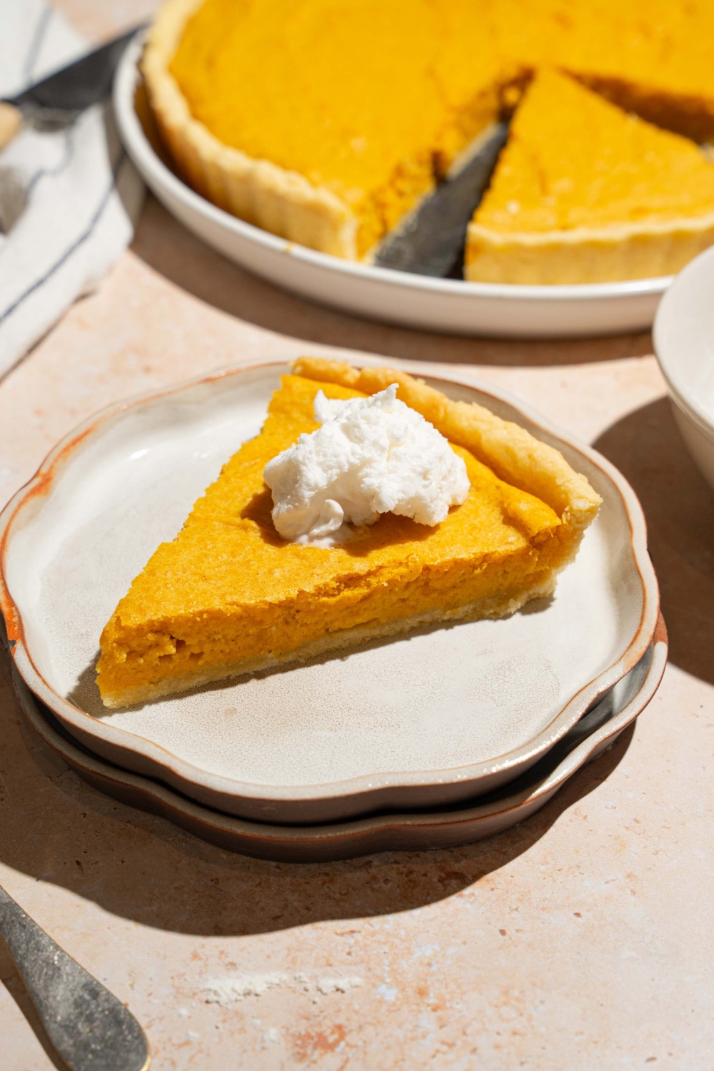 A plate with a slice of cream cheese pumpkin pie topped with a dollop of whipped cream. The plate is on a tan counter with a whole pie and white striped napkin.