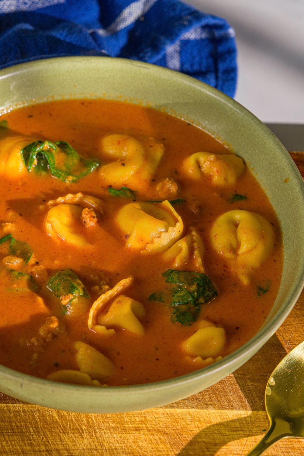 A bowl of creamy tomato tortellini soup. The bowl is on a wooden board with a spoon and a blue cloth napkin.
