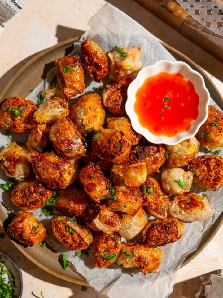 A plate lined with parchment paper with crispy rice paper chicken bites garnished with fresh parsley. There is a bowl of dipping sauce on the plate. The plate is on a tan counter.
