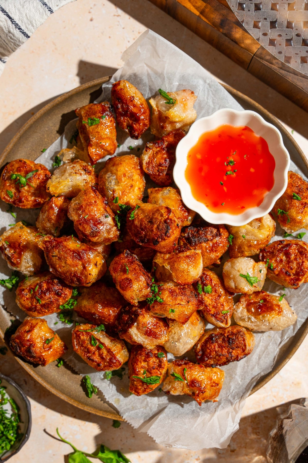 A plate lined with parchment paper with crispy rice paper chicken bites garnished with fresh parsley. There is a bowl of dipping sauce on the plate. The plate is on a tan counter.