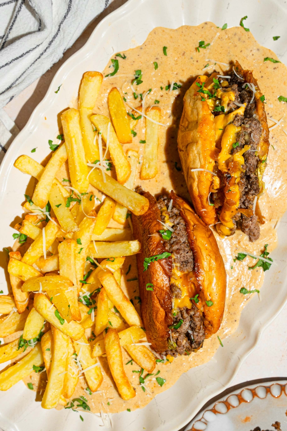 A cheeseburger that is cut in half with the 2/2 faced down in a creamy garlic Parmesan sauce that is on a white plate. Next to the burgers to the left are french fries also in the garlic Parmesan sauce.