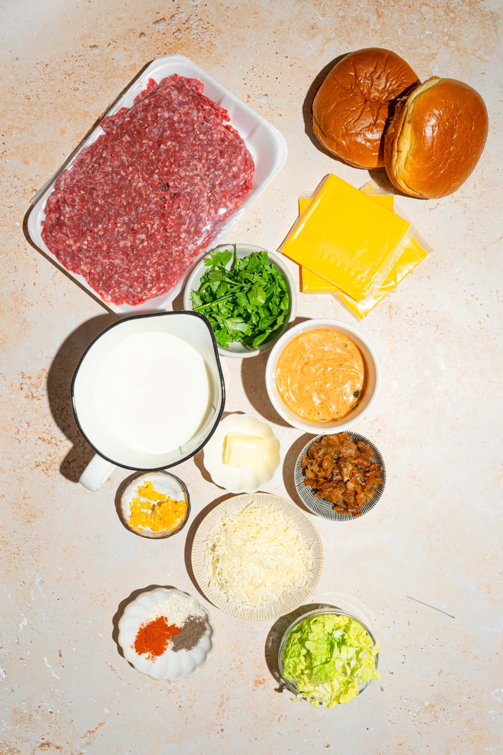 Ground beef in a package, two burger buns, two slices of American cheese, a picture of half-and-half, a bowl of burger sauce, a bowl of caramelized, onions, a bowl of garlic powder, onion powder, and black pepper, a bowl of lettuce, and a bowl of grated Parmesan cheese on a white counter.