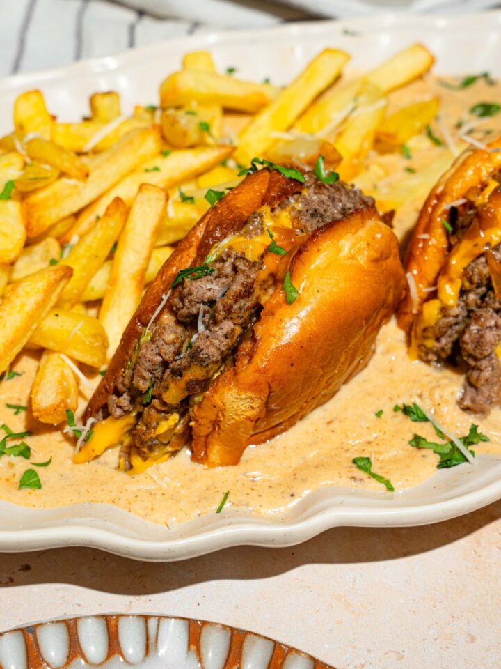 A half of a cheeseburger that is faced down in a creamy garlic Parmesan sauce on a white plate. Behind the burger are french fries and next to it is part of the other half of the burger.