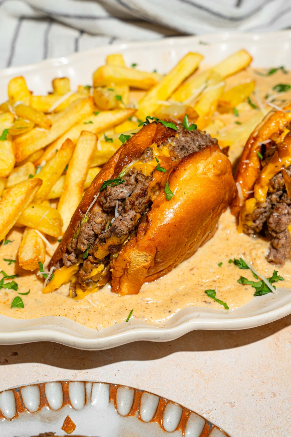 A half of a cheeseburger that is faced down in a creamy garlic Parmesan sauce on a white plate. Behind the burger are french fries and next to it is part of the other half of the burger.