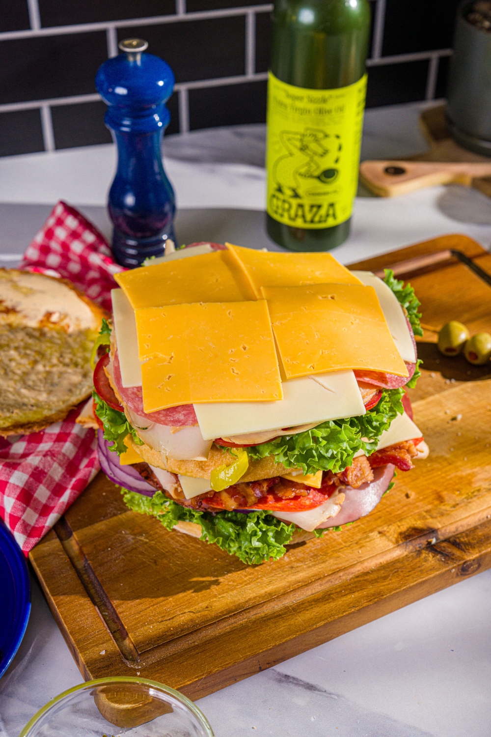 Two layers of cheddar cheese, swiss cheese, lettuce, tomato, onion, deli meats, and bread all stacked on a wood cutting board.