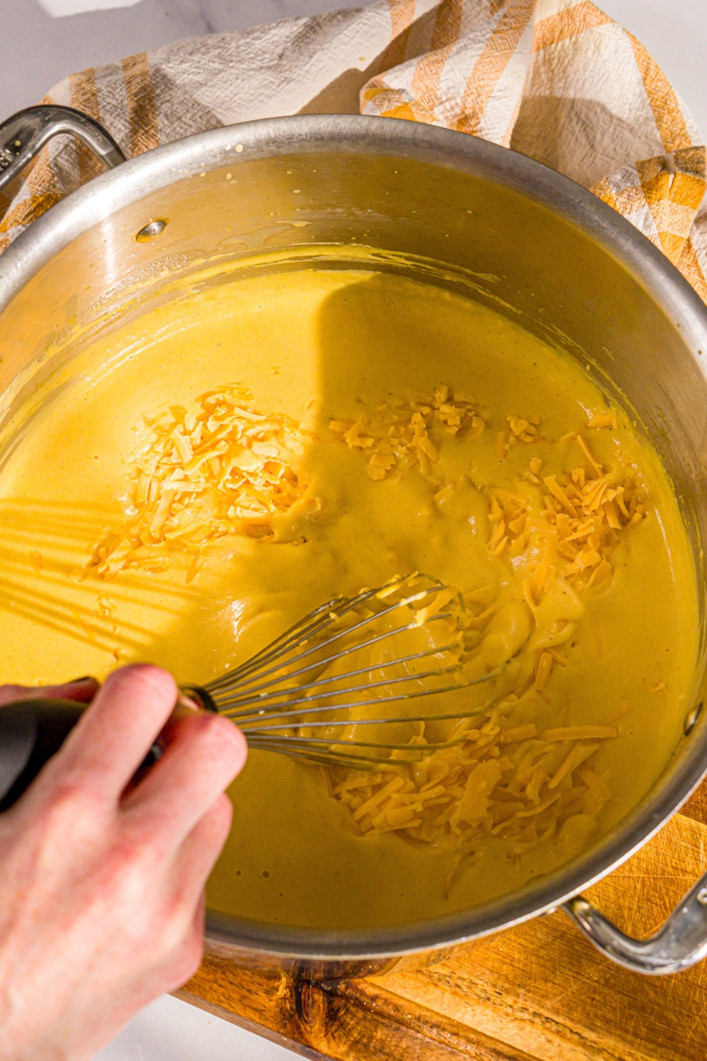A hand holding a whisk, whisking shredded cheddar cheese into a mixture of roasted butternut squash puree in a large pot.