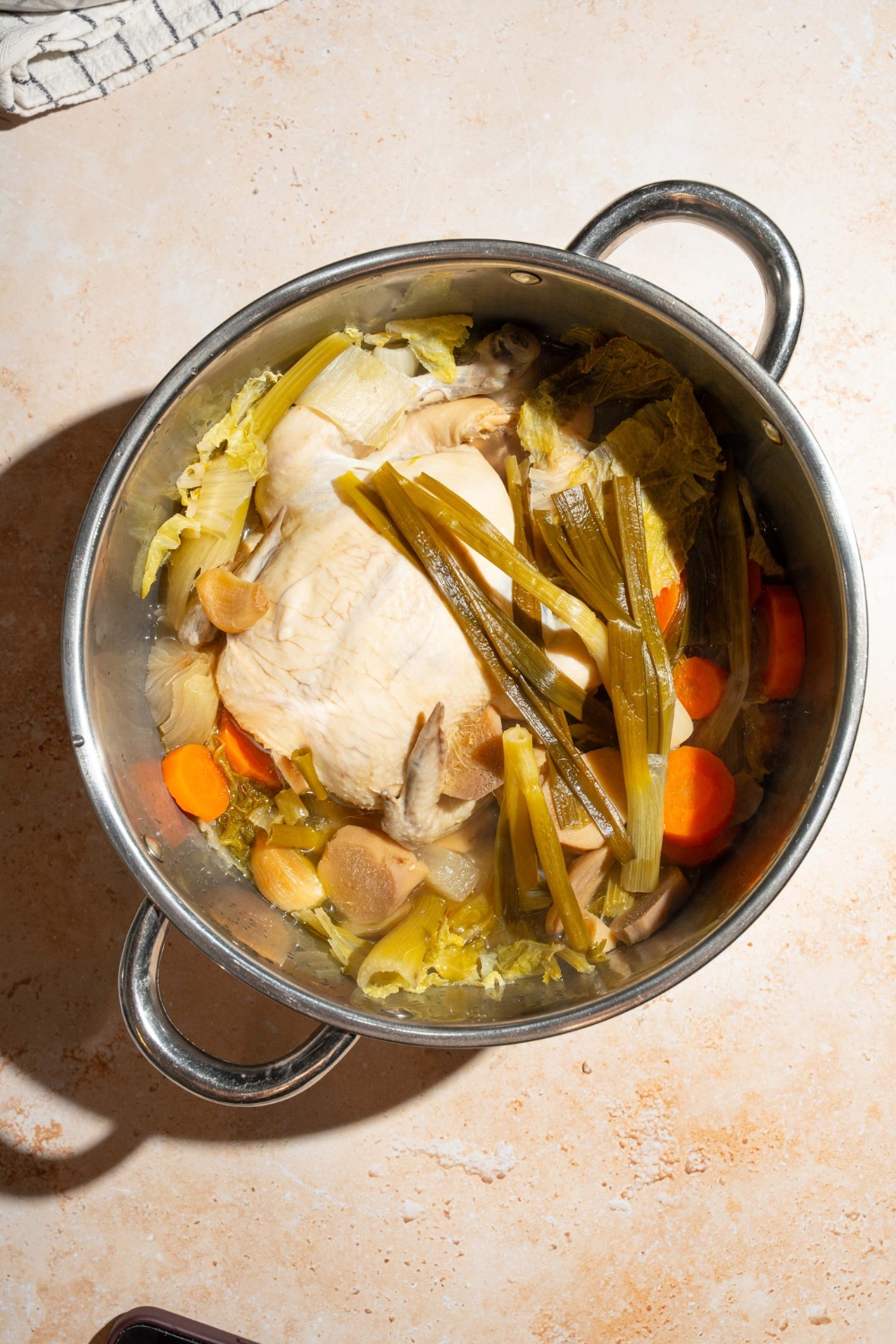 Cooked scallions on top of a whole chicken, with carrots, celery, garlic and cabbage in a large pot for a no water chicken soup.