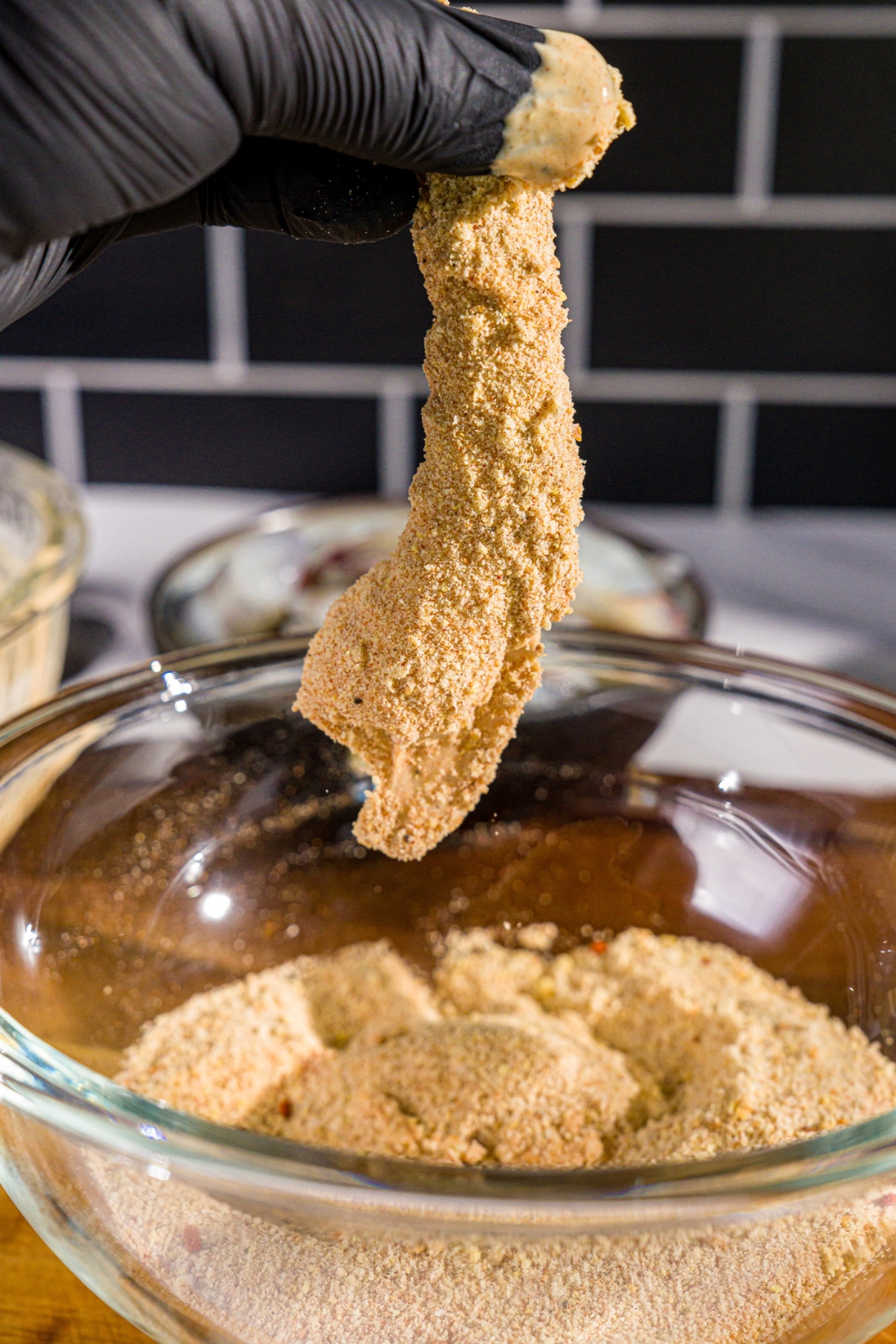 A hand dipping an uncooked battered prawn into a bowl of seasoned breadcrumbs. The bowl is on a wooden board.