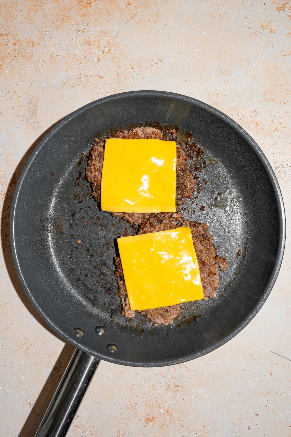 American cheese slices on top of two burger patties cooking in a skillet.