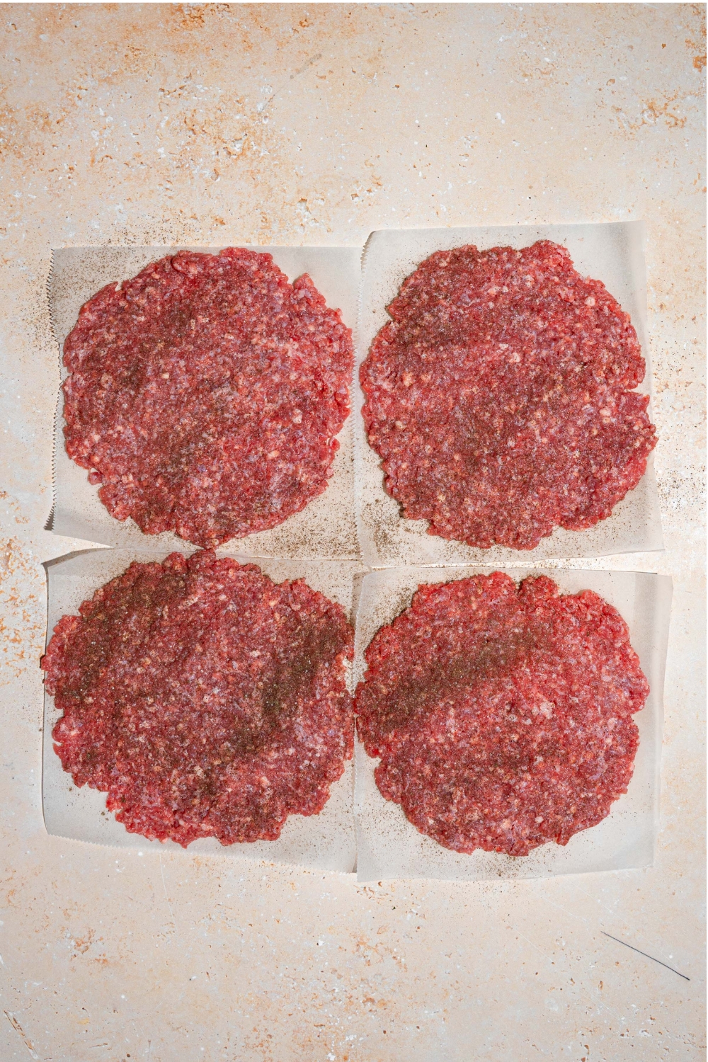 Four raw burger patties with salt and pepper on top sit next to one another square on top of a paper sheet on a white counter.