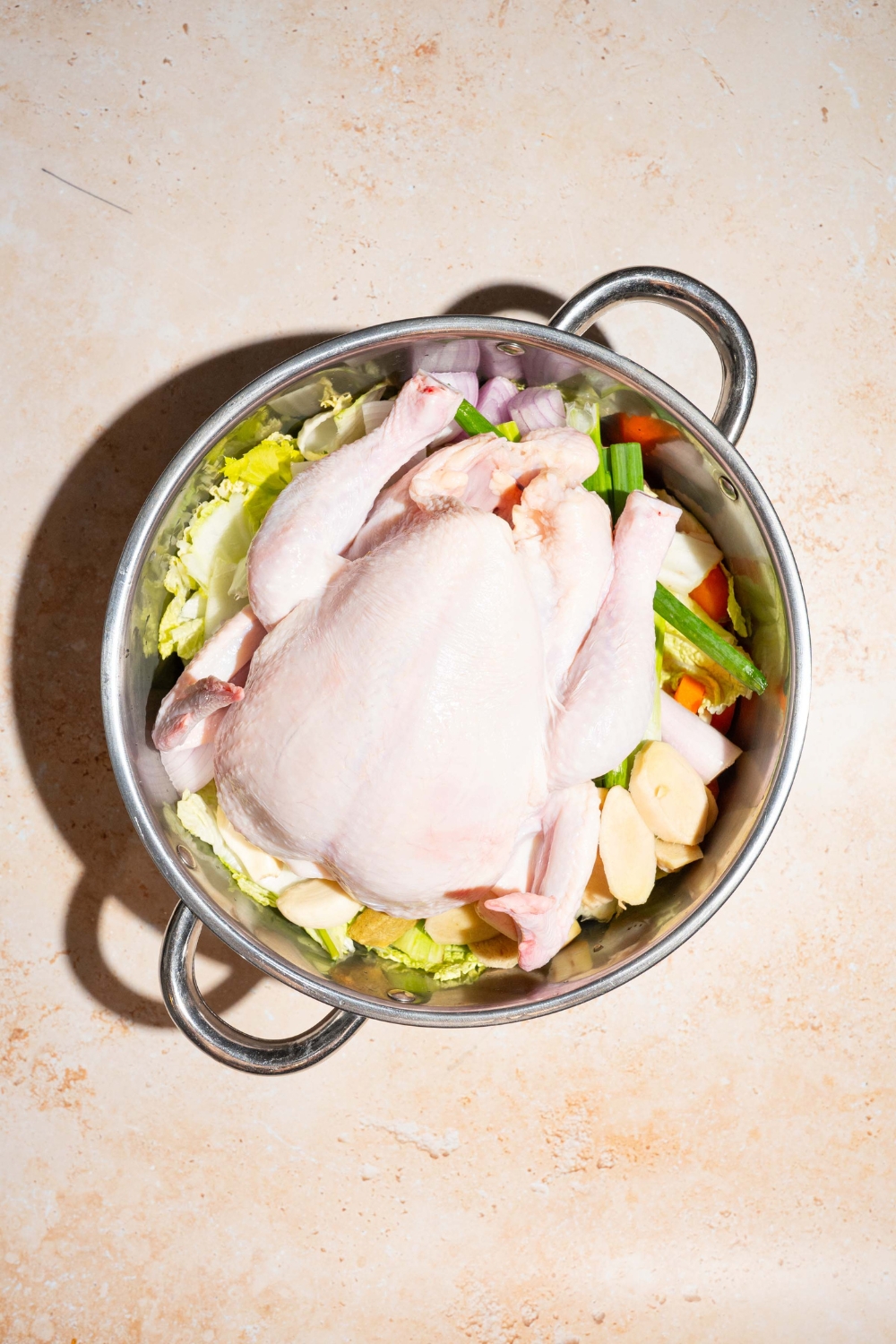 A whole chicken on top of chopped carrots, onions, scallions, celery, and garlic in a large pot.
