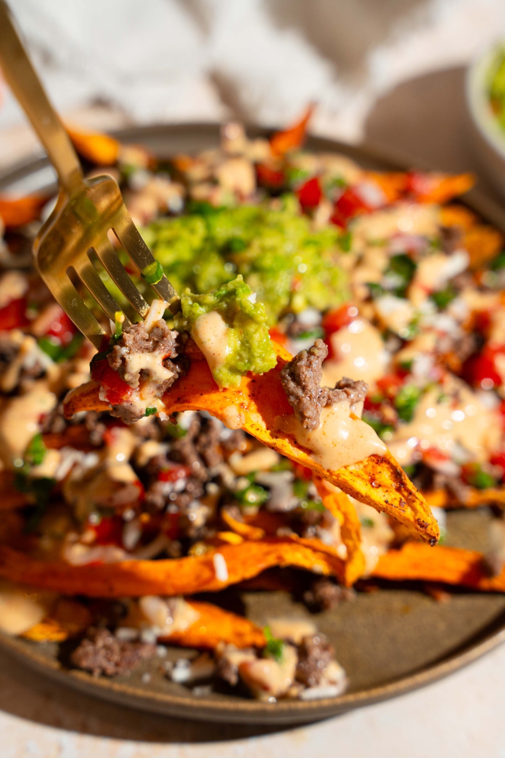 A close up of a fork with a bite of sweet potato loaded fries with ground beef, chipotle ranch dressing, pico de gallo, shredded cheese, and guacamole. There is a plate of loaded fries blurred in the background.