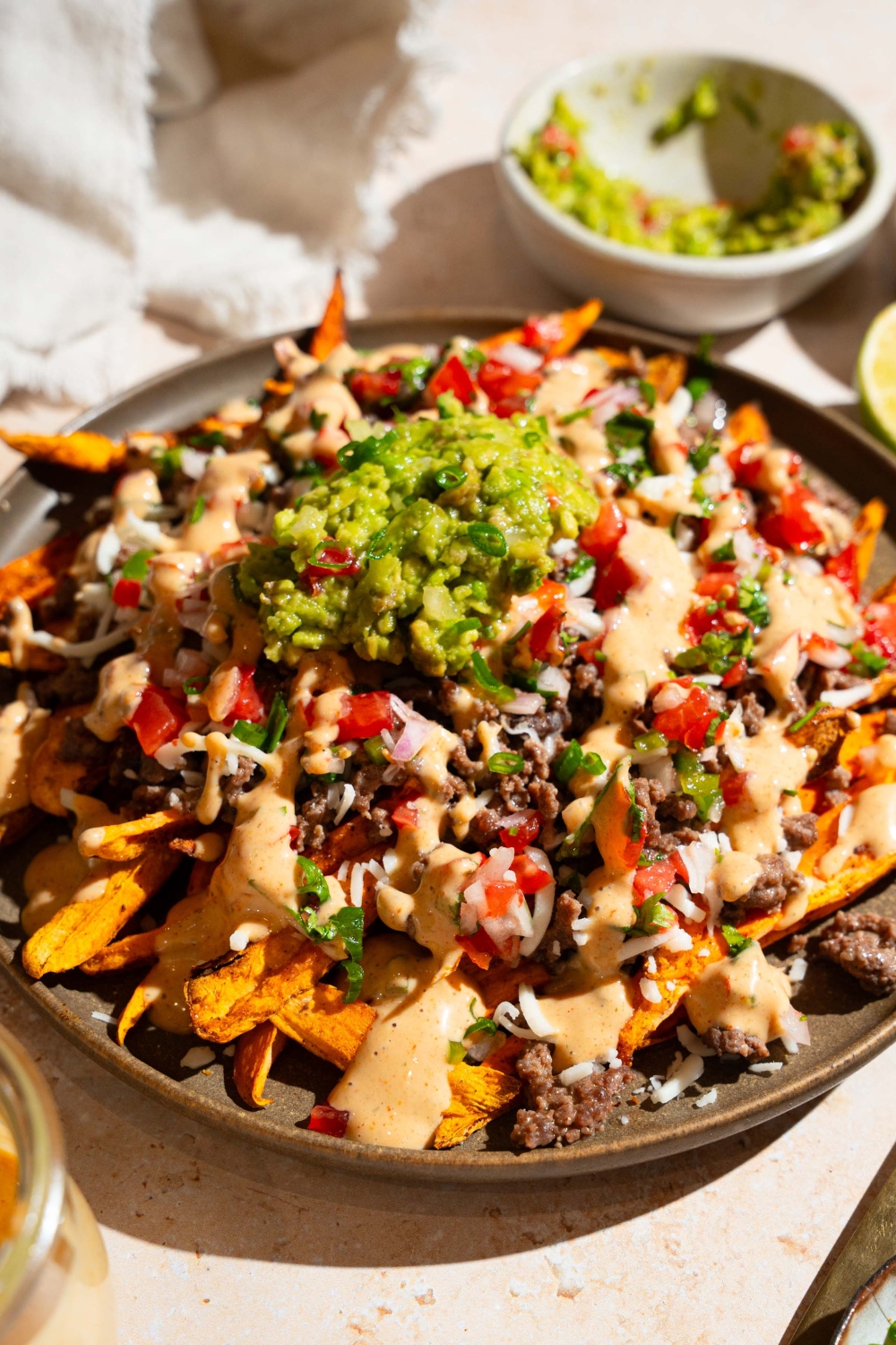 A plate of loaded sweet potato fries topped with ground beef, chipotle ranch, pico de gallo, shredded cheese, and guacamole. The plate is on a tan counter with small bowls of toppings.