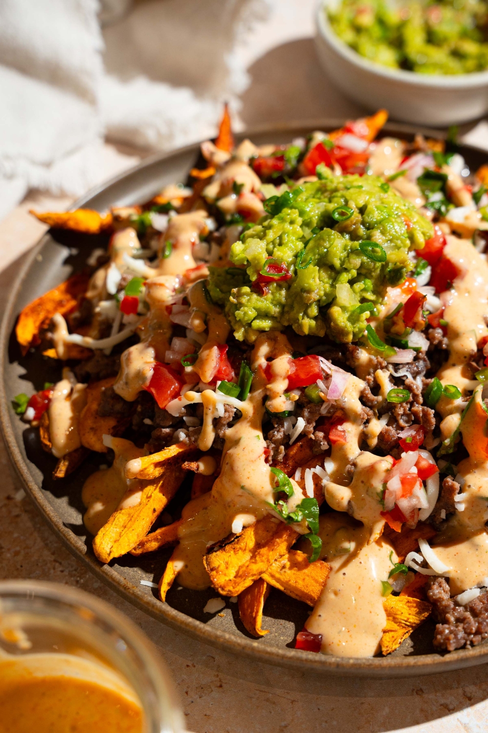 A plate of loaded sweet potato fries topped with ground beef, chipotle ranch, pico de gallo, shredded cheese, and guacamole. The plate is on a tan counter with small bowls of toppings.