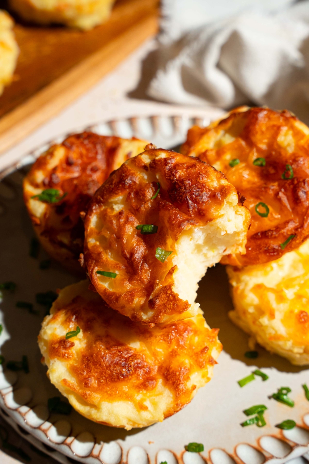 A plate with several mashed potato cups garnished with sliced green onion. One of the cups has a bite removed. The plate is on a tan counter with a white cloth napkin.
