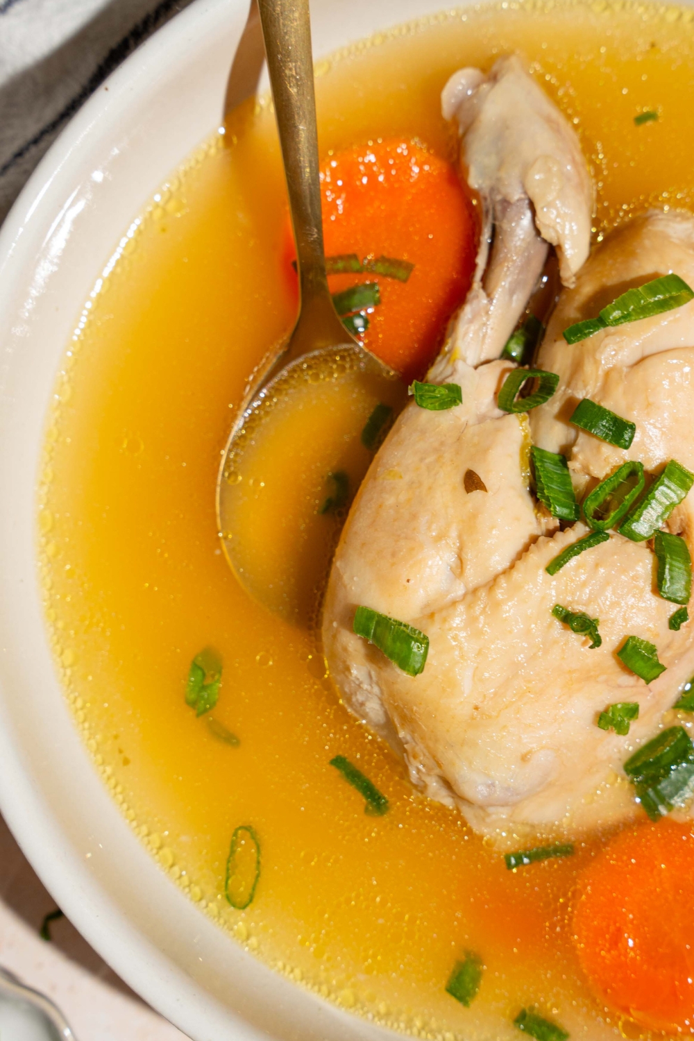Part of a white bowl that is filled with no water chicken soup. The bowl has part of a whole chicken and some sliced carrots in it.
