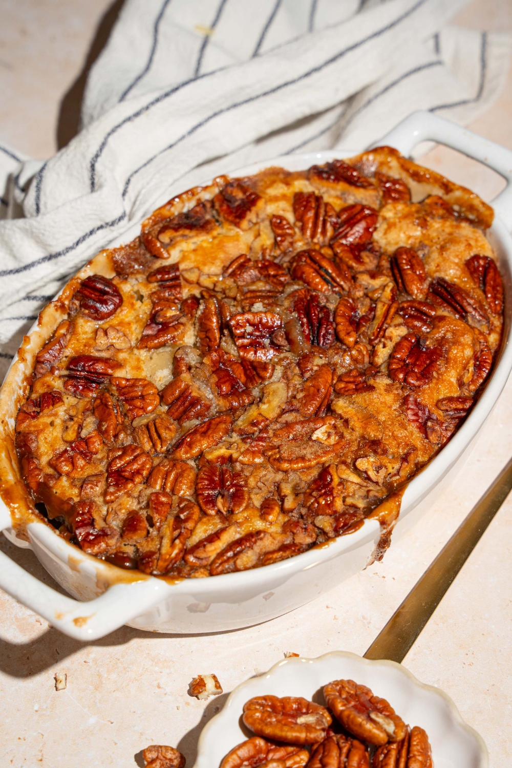A casserole dish with baked pecan pie cobbler. The dish is on a tan counter with a white striped napkin and small plate of pecans.