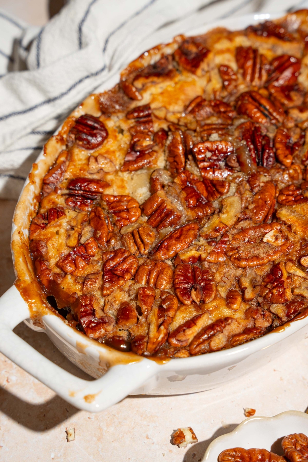 A casserole dish with baked pecan pie cobbler. The dish is on a tan counter with a white striped napkin and small plate of pecans.