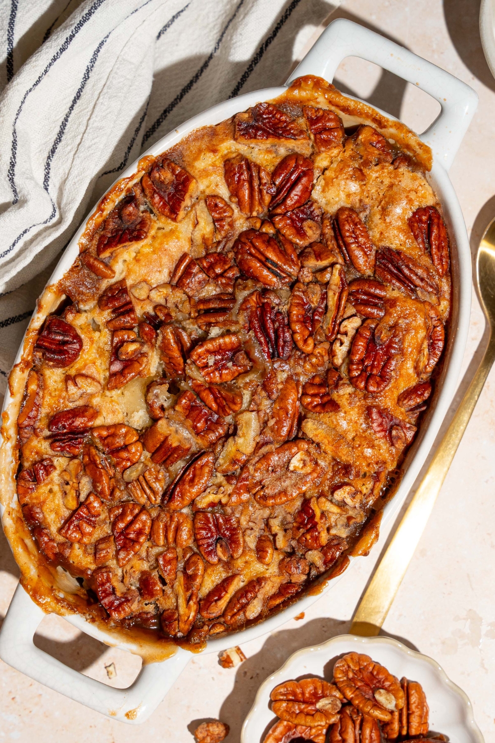 A casserole dish with baked pecan pie cobbler. The dish is on a tan counter with a white striped napkin and small plate of pecans.