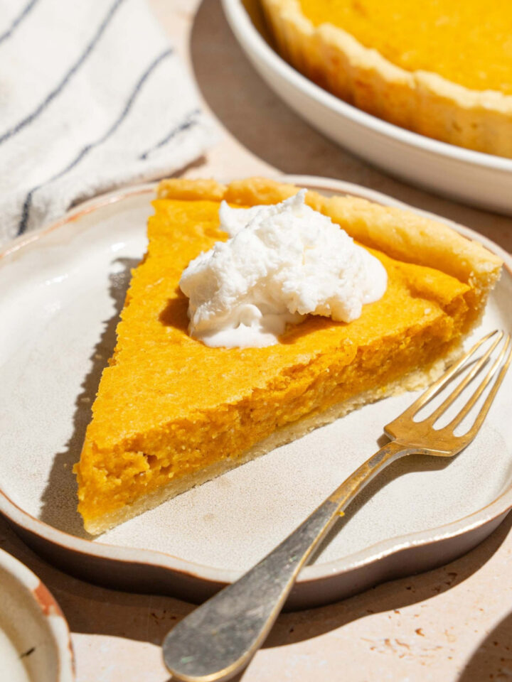 A slice of pumpkin cream cheese pie with a dollop of whipped cream on top on a white plate with a fork. Part of the whole pumpkin cream cheese pie is behind it.