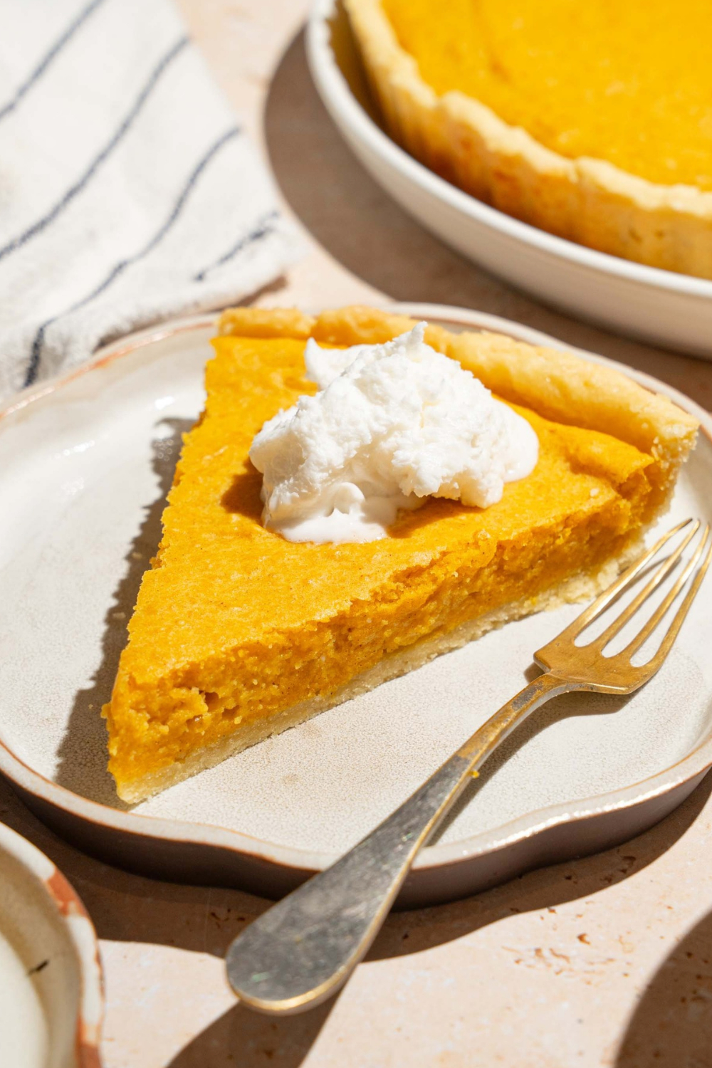 A slice of pumpkin cream cheese pie with a dollop of whipped cream on top on a white plate with a fork. Part of the whole pumpkin cream cheese pie is behind it.