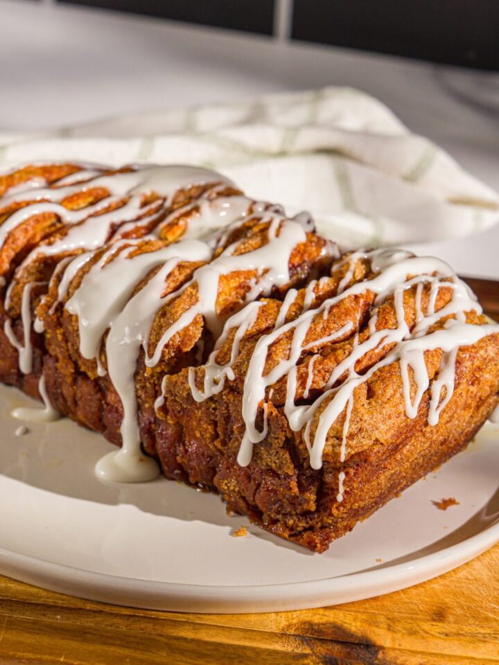 A plate of pumpkin pull apart bread loaf drizzled with glaze. The plate is on a wooden board with a white cloth napkin.