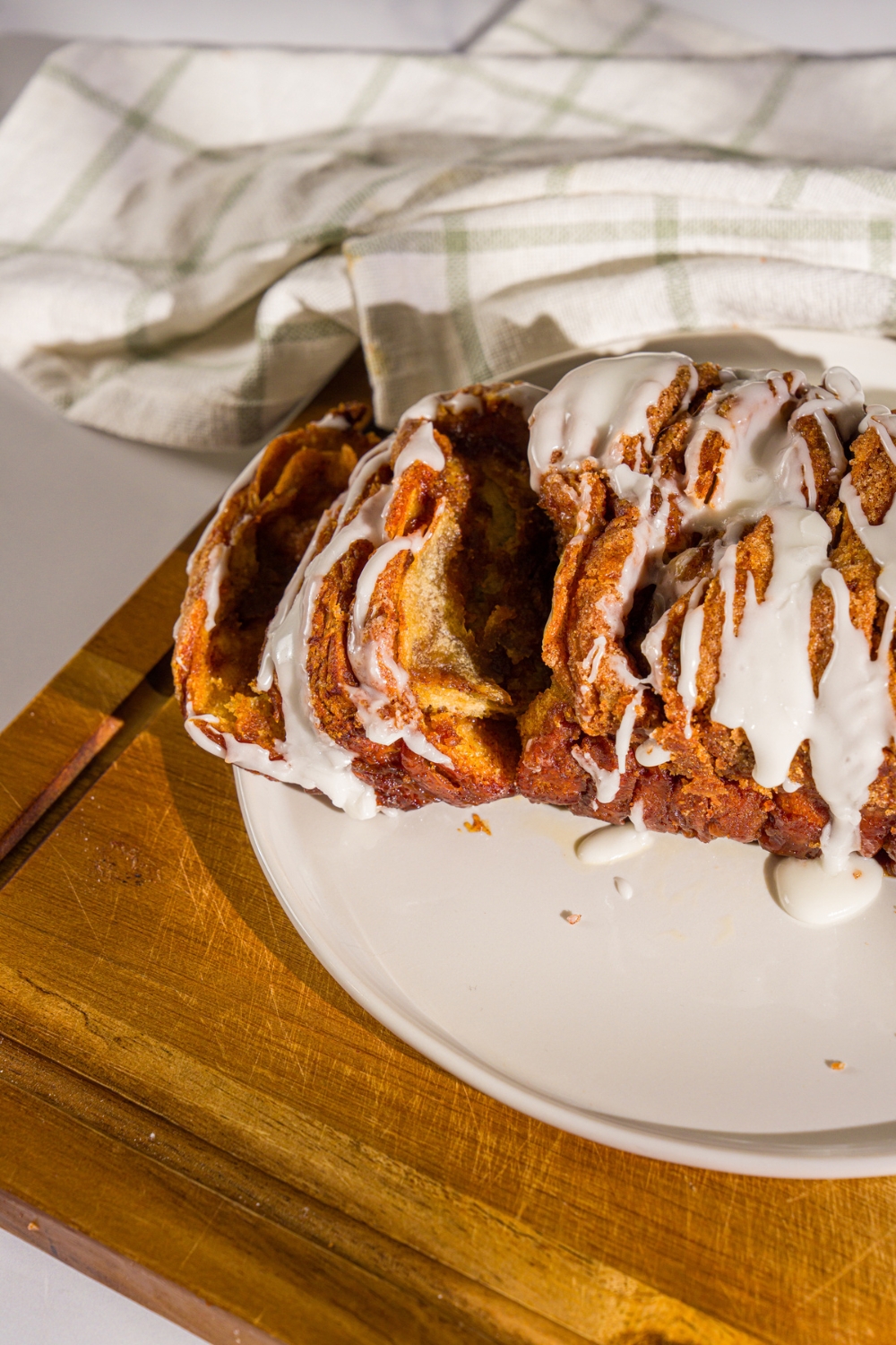 A plate of pumpkin pull apart bread loaf drizzled with glaze. Two slices have been cut from the loaf. The plate is on a wooden board with a white cloth napkin.