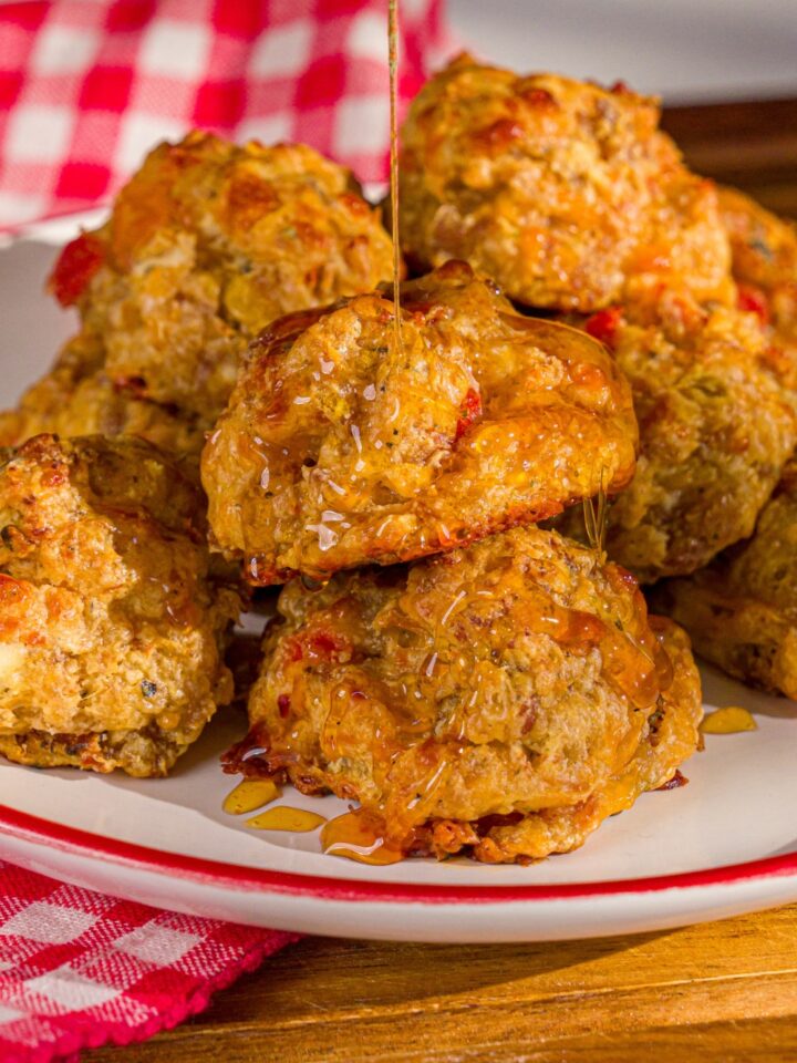 A plate of cheddar bay biscuit sausage balls being drizzled with hot honey. The plate is on a wooden board with a red checkered napkin.