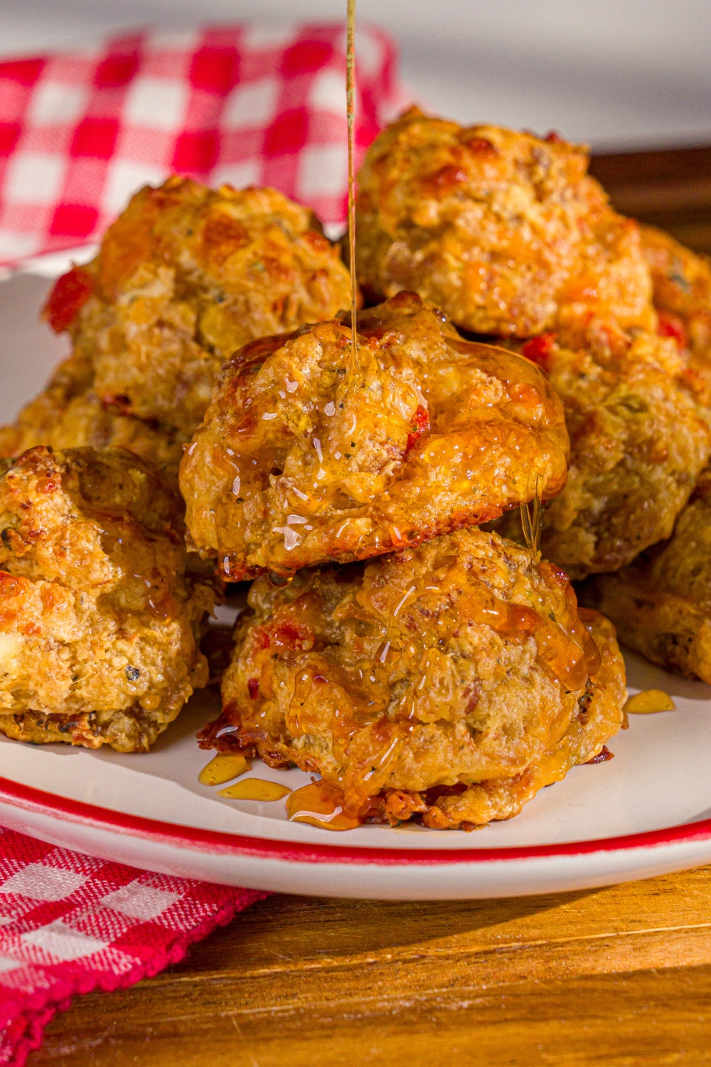A plate of cheddar bay biscuit sausage balls being drizzled with hot honey. The plate is on a wooden board with a red checkered napkin.
