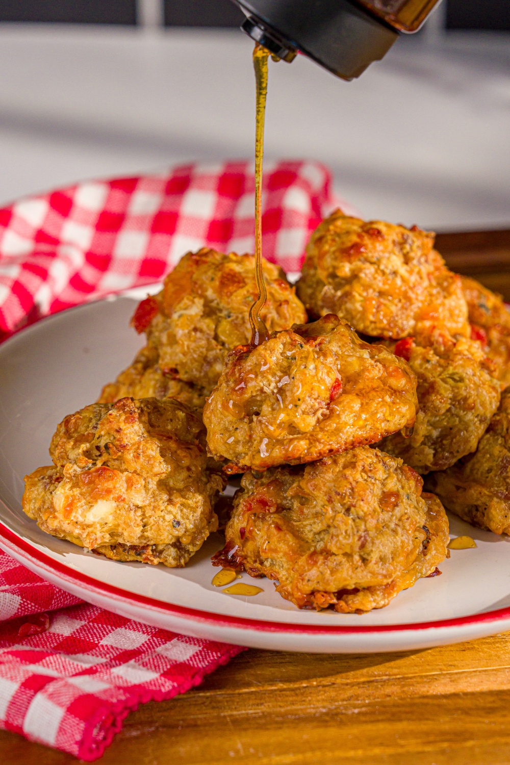 A plate of cheddar bay biscuit sausage balls being drizzled with hot honey. The plate is on a wooden board with a red checkered napkin.