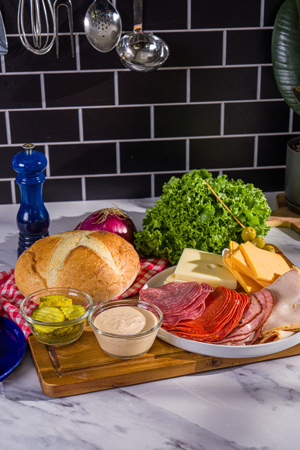 An Italian bread round, a bowl of pickles, a head of lettuce, a cup of chipotle mayo, a plate with sliced turkey, ham, salami, pepperoni, swiss, and cheddar cheese all on a wood cutting board, all for a scooby doo sandwich.