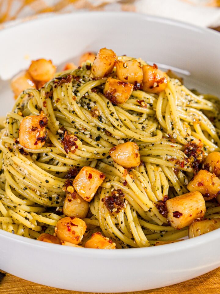 A bowl of seaweed pasta topped with seared scallops and chili crisp. The bowl is on a wooden board.