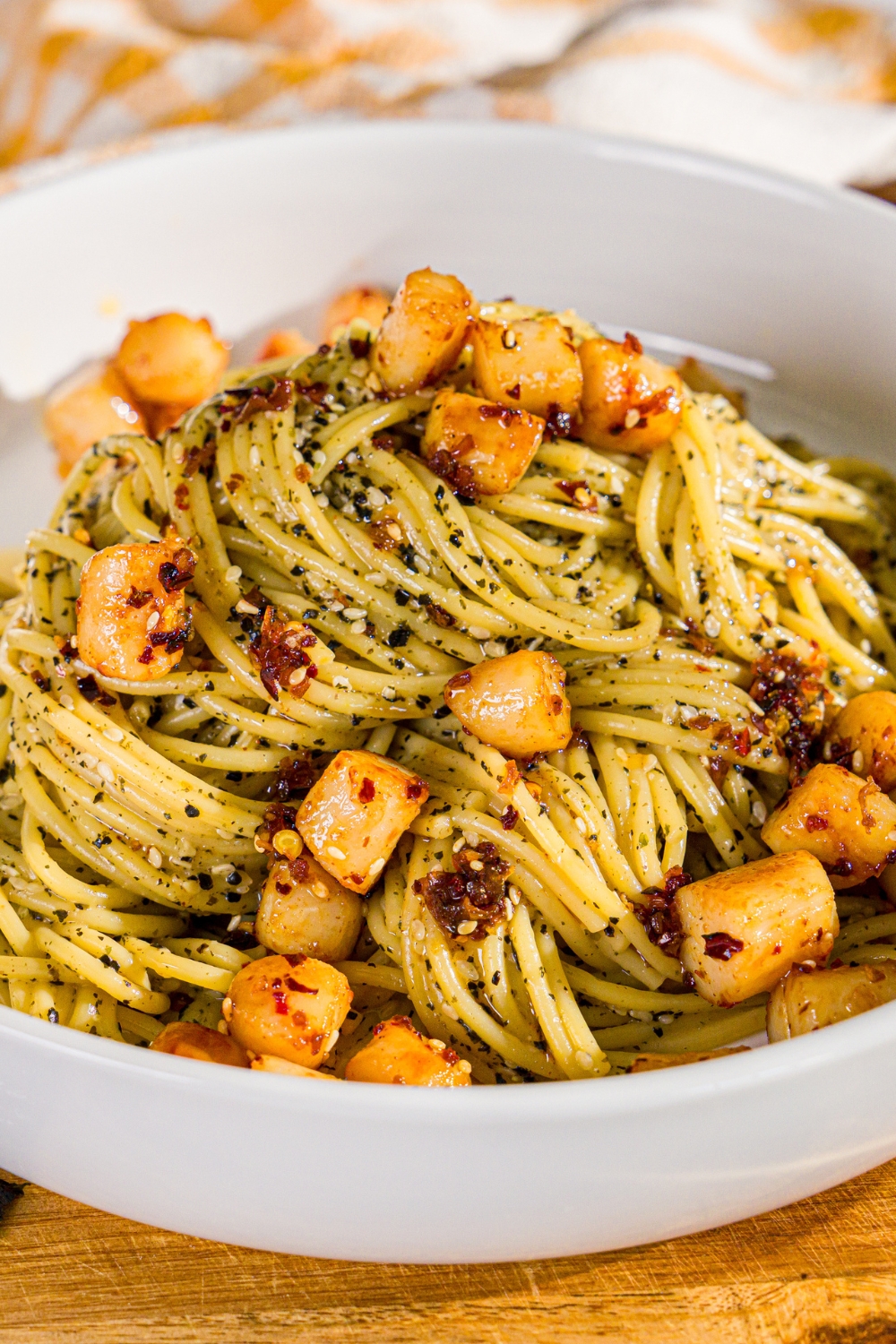 A bowl of seaweed pasta topped with seared scallops and chili crisp. The bowl is on a wooden board.