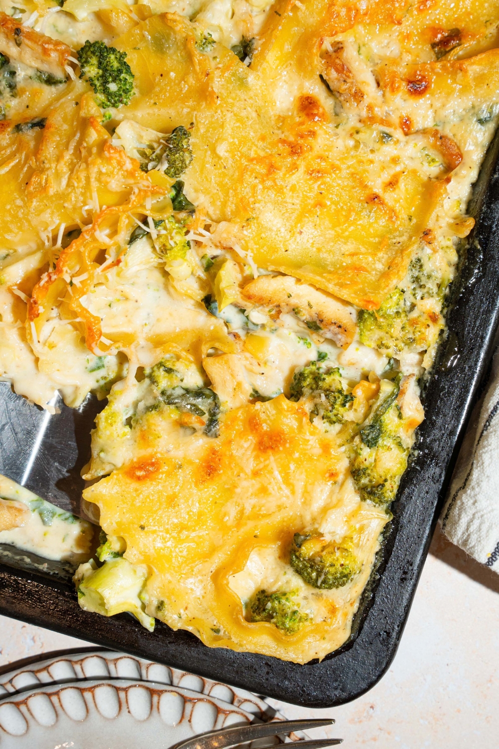 A sheet pan with baked chicken broccoli lasagna. There is a spatula taking a slice from the pan. The pan is on a tan counter.