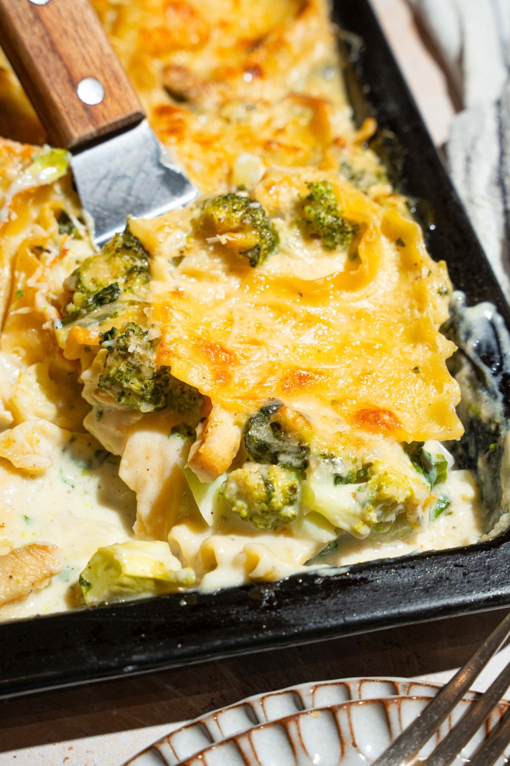 A sheet pan with baked chicken broccoli lasagna. There is a spatula taking a slice from the pan. The pan is on a tan counter.