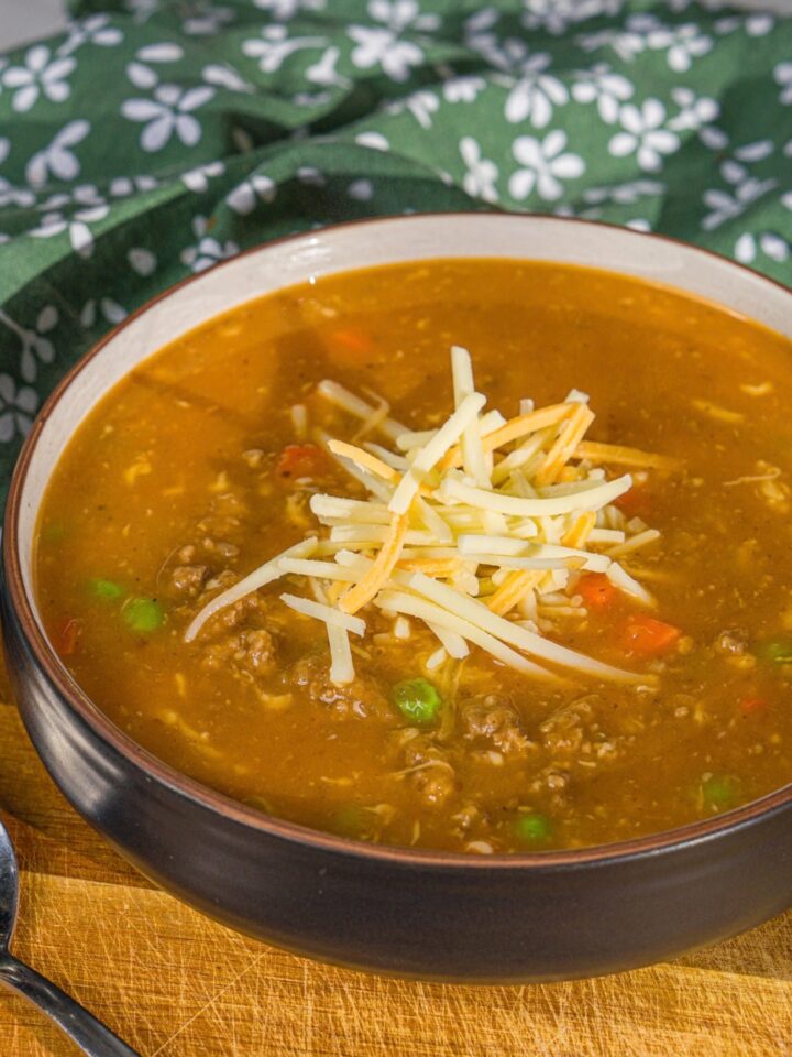 A bowl of shepherd's pie soup topped with shredded cheese. The bowl is on a wooden board with a spoon and green cloth napkin.