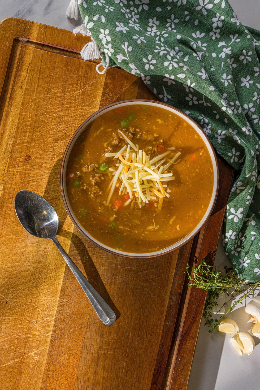A bowl of shepherd's pie soup topped with shredded cheese. The bowl is on a wooden board with a spoon and green cloth napkin.