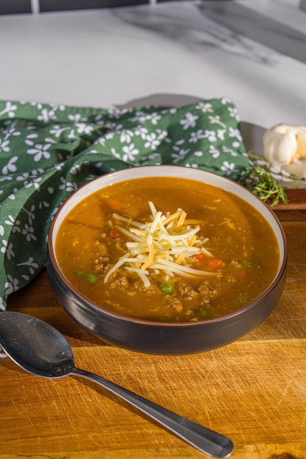 A bowl of shepherd's pie soup topped with shredded cheese. The bowl is on a wooden board with a spoon and green cloth napkin.
