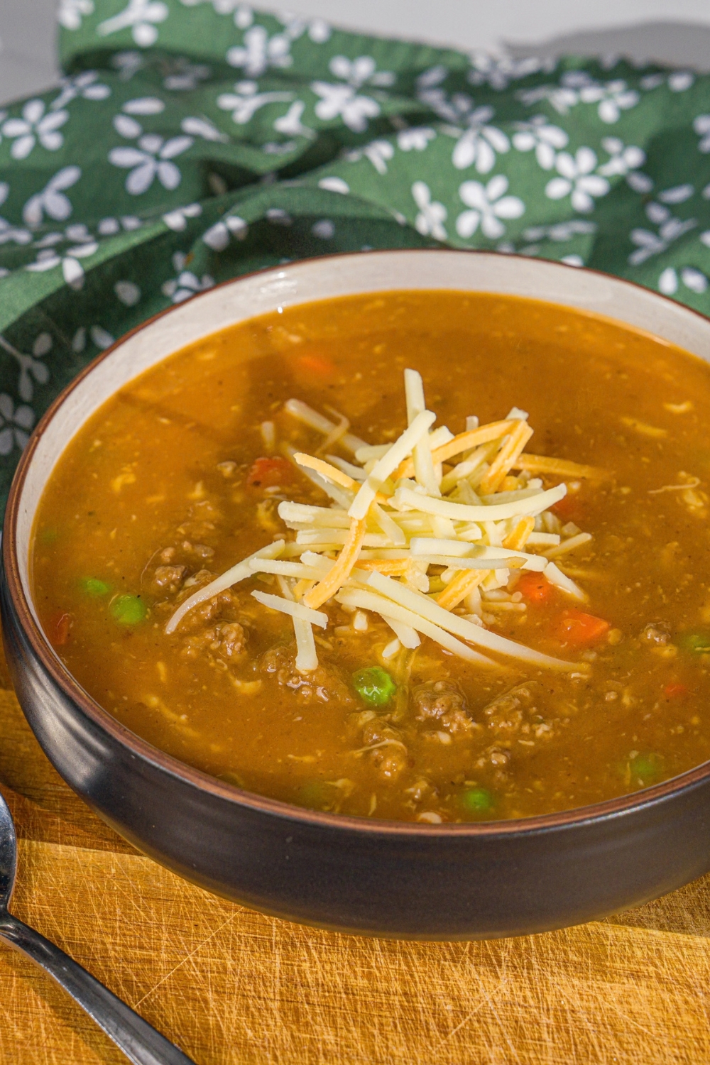 A bowl of shepherd's pie soup topped with shredded cheese. The bowl is on a wooden board with a spoon and green cloth napkin.