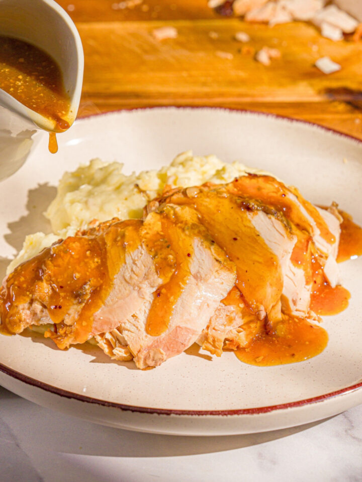 A few slices of slow cooker boneless turkey breast with gravy on top on a white plate with mashed potatoes behind them. A white pitcher filled with the gravy is being held to the left of the plate.
