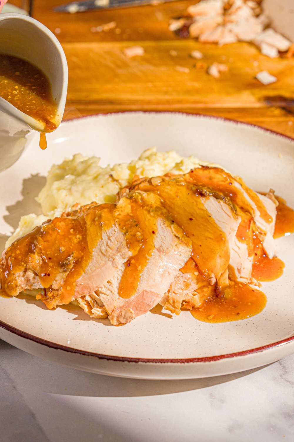 A few slices of slow cooker boneless turkey breast with gravy on top on a white plate with mashed potatoes behind them. A white pitcher filled with the gravy is being held to the left of the plate.