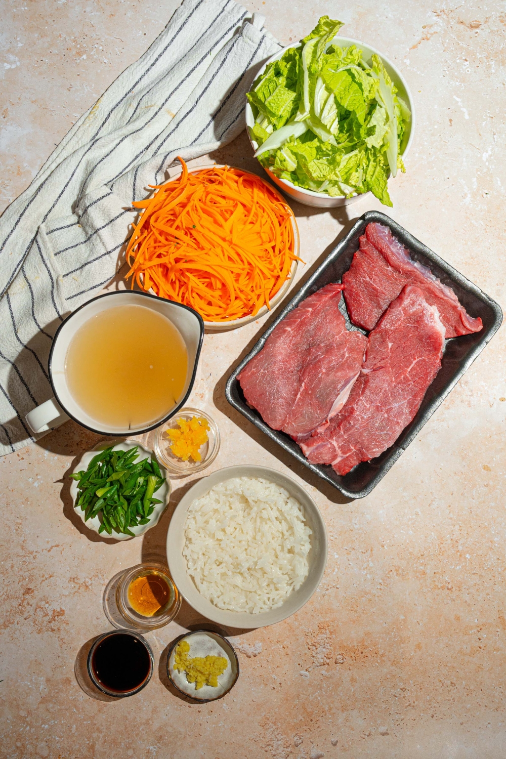 A bowl of shredded carrots, bok choy, a pitcher of broth, a bowl of white rice, and two sirloin steaks in a package all on a white counter.