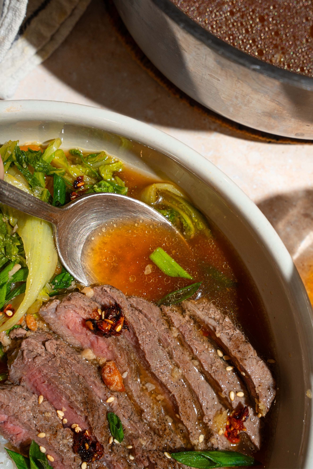 A spoon submerged in broth that is in a white bowl with sliced steak and shredded bok choy.