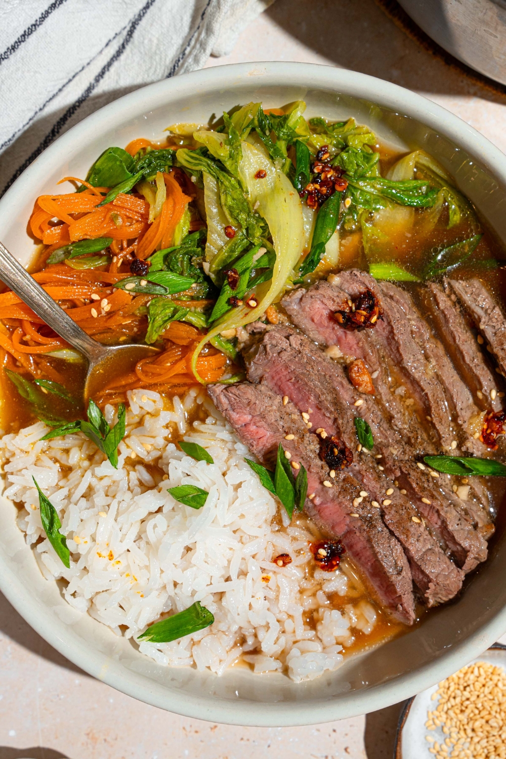A steak and rice broth bowl with jasmine rice, sliced sirloin steak, and shredded carrots and bok choy submerged in broth in a white bowl.