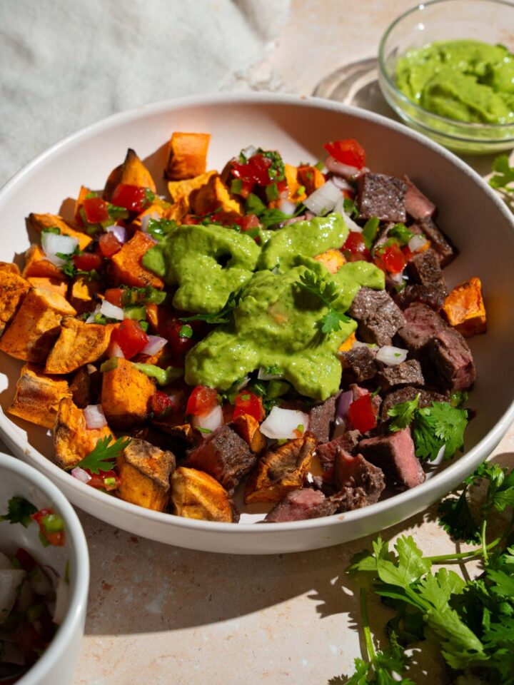 A sweet potato steak bowl topped with pico de gallo and avocado cilantro lime dressing. The bowl is on a tan counter with fresh cilantro with a small bowl of dressing.