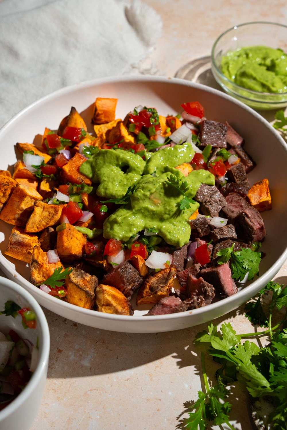 A sweet potato steak bowl topped with pico de gallo and avocado cilantro lime dressing. The bowl is on a tan counter with fresh cilantro with a small bowl of dressing.
